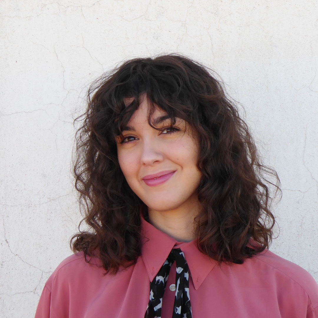 Portrait of Miriam smiling, she has dark curly hair and is wearing a pink blouse.